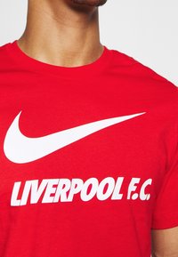 Camiseta de algodón roja con el logo de Nike en blanco y texto en negrita que dice "LIVERPOOL F.C." en el frente. Mangas cortas, cuello redondo.