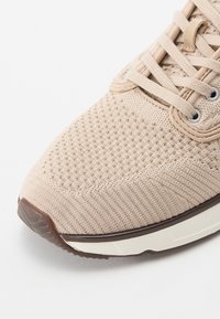 Beige sneakers med tekstureret stofoverdel, der har et vævet mønster, rund tå og en kontrast brun gummisål. Snørebånd og øjer synlige.