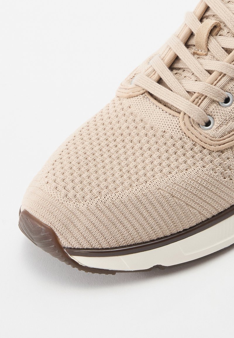 Baskets beiges avec une tige en tissu texturé, présentant un motif tissé, un bout rond et une semelle en caoutchouc marron contrastante. Lacets et œillets visibles.
