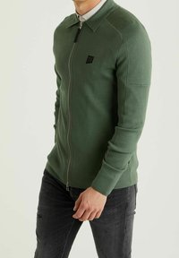 Grön zip-up tröja med krage, tillverkad av texturerat tyg. Har en liten svart logotyp på bröstet och ribbade muddarna, bärs tillsammans med mörka jeans.