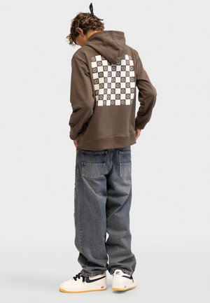 Persona con capelli ricci che indossa una felpa marrone con cappuccio con una scacchiera bianca e faccine sorridenti sulla schiena, jeans larghi e sneakers Nike bianche.