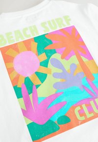 T-shirt blanc avec un design graphique coloré, comprenant un soleil rose, un arrière-plan vert et bleu, et des plantes tropicales en rose, violet et orange.