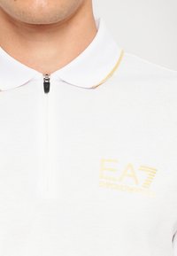 Balta polo marškinėliai su geltonu akcentu ant apykaklės, turintys pusiau užtrauktuką ir išsiuvinėtą EA7 Emporio Armani logotipą krūtinėje.