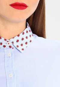 Světle modrá košile s vzorovaným límcem, na kterém jsou červené berušky na bílém a modrém pruhovaném pozadí. Viditelné jsou dvě bílé knoflíky.