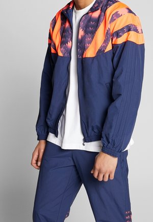 Man draagt een marineblauw trainingspak met feloranje en paarse geometrische patronen op de schouders en bovenborst van het jack.