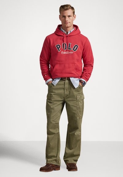 Muškarac u crvenoj polo majici Ralph Lauren preko plave košulje, maslinasto zelenim cargo hlačama i smeđim cipelama, stoji s rukama u džepovima.