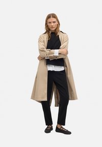 Trench beige sobre un suéter negro, camisa blanca y pantalones negros. Zapatos planos negros con correas. El modelo está de pie con los brazos cruzados.