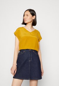 Top amarelo mostarda em V com mangas curtas, combinado com uma saia de ganga escura de cintura alta com costuras em contraste e bolsos frontais.