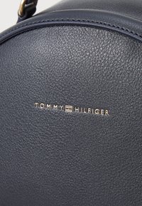 Tumesinine tekstuuriga nahk kott ümarate vormidega, mille esiküljel on kuldne reljeefne "TOMMY HILFIGER" logo.