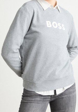 Personne portant un sweat-shirt gris avec le texte "BOSS" sur une chemise blanche à col et un jean gris, debout les mains derrière le dos.