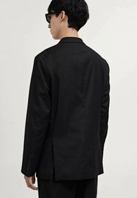 Black tailored jacket made of smooth fabric, featuring a single vent at the back and buttoned cuffs. Worn with dark sunglasses.
