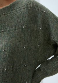 Mörkgrön stickad tröja med en mjuk textur, med horisontella ränder och dekorativa silverstift över hela.