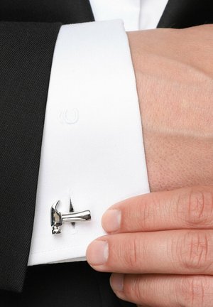 Srebrny spinkowy w kształcie młotka, przypięty do białego mankietu, w zestawieniu z czarną marynarką. Zbliżenie pokazujące ułożenie ręki.