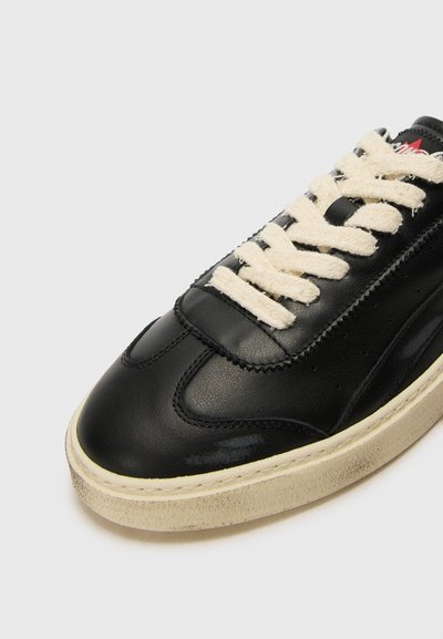 Zapatilla de cuero negro con punta redondeada, cordones color crema y una suela texturizada. Presenta costuras a lo largo de los lados y un branding minimalista.