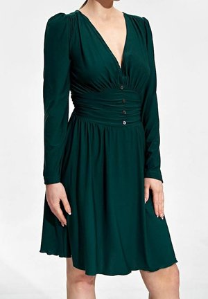 Femme portant une robe vert foncé à manches longues avec un décolleté en V profond, une taille froncée ornée de boutons et une jupe évasée jusqu'aux genoux.