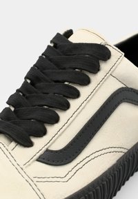 Zapatilla de ante blanca con cordones negros, que cuenta con una prominente franja lateral de goma negra y suela texturizada negra. Diseño minimalista con colores contrastantes.
