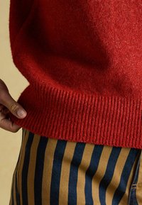 Roter Wollpullover mit geripptem Saum, kombiniert mit blau- und senfgestreiften Hosen. Nahaufnahme zeigt die Textur und Details des Stoffes.