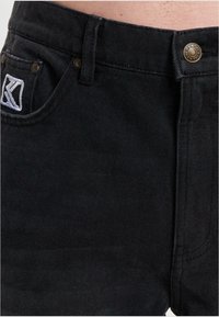 Jeans de denim negro con un diseño ajustado, costuras visibles, un botón en la parte frontal y una etiqueta de la marca en el bolsillo trasero. Textura suave.