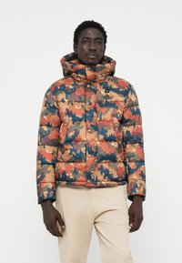 Veste camouflage rembourrée avec un col haut, présentant un mélange de couleurs orange, bleu et beige, des boutons-pression, et une texture lisse et brillante.