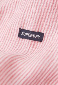 Tejido a rayas rojas y blancas con una textura suave. Incluye una etiqueta azul marino con letras blancas que dice "SUPERDRY".