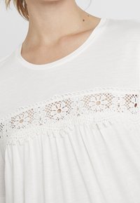 Vit topp med rund halsringning, detaljerad spetskant med blommönster och plisserad nedre del. Mjuk, lätt tygkvalitet.