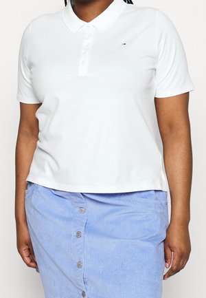 Polo blanc à manches courtes avec col. Présente un petit logo sur la poitrine. Associé à une jupe en velours côtelé bleue avec des boutons.