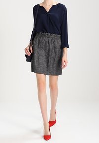 Žena oblečená v námořnickém modrém topu, šedé texturované sukni, červených špičatých podpatcích a nesoucí černou kabelku přes rameno, stojící na bílém pozadí.