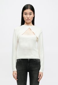 Λευκό πλεκτό πουλόβερ με μακριά μανίκια, διαθέτει τριγωνική κοπή στο στήθος, κουμπί κλεισίματος και ριχτό τελείωμα. Φοριέται πάνω από ένα εφαρμοστό τοπ.