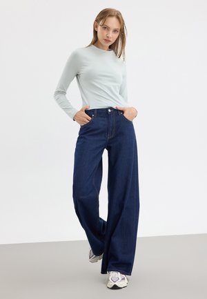 Vaaleansininen pitkähihaisen paidan, tummansiniset väljät farkut etutaskuilla, päällä valkoiset ja violetit lenkkarit, vaaleanharmaalla lattialla.