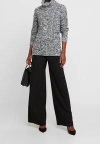 Schwarzer und weißer melierter Strickpullover mit einem Schalkragen, kombiniert mit weit geschnittenen schwarzen Hosen. Außerdem ein kleiner schwarzer Handtasche und hohe Absätze.