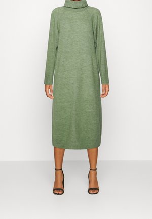 Robe en maille verte mi-longue pour femme avec col roulé et manches longues, associée à des sandales à talons hauts noires avec bride à la cheville, debout sur fond blanc.