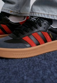 Fekete és szürke sportcipők piros velúr csíkokkal, gumitalppal és fekete fűzővel, a sarkán "Samba" márkázással.