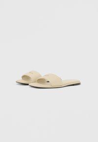Hellebeige Rutsch-Sandalen mit einem breiten Oberband, flacher Sohle und subtilen Nähten entlang der Kanten. Glatte, matte Textur.