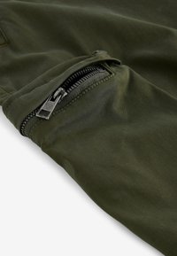 Pantalonii de culorile verzi măslinii au un buzunar cu fermoar de culoare neagră și căptușeală turcoaz. Materialul are o textură netedă cu un ușor luciu.