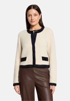 Vrouw draagt een crèmekleurig wollen vest met zwarte bies, zwarte knopen en voorzakken, gecombineerd met een hooggesneden bruine leren broek.