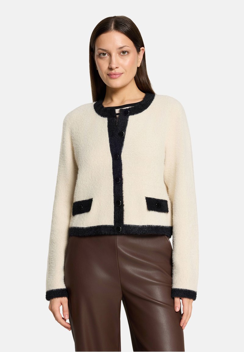 Femme portant un cardigan en laine crème avec bordures noires, boutons noirs et poches avant, assorti à un pantalon en cuir marron taille haute.