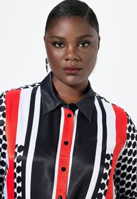 Camisa de satén multicolor con rayas verticales en rojo, negro, blanco y patrones geométricos, con un collar puntual y botones negros.
