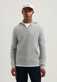 Man in een lichtgrijze geribbelde sweater met kwartrits, een wit T-shirt eronder, marineblauwe broek en een witte pet met blauwe tekst "NO STREZZ CLUB".