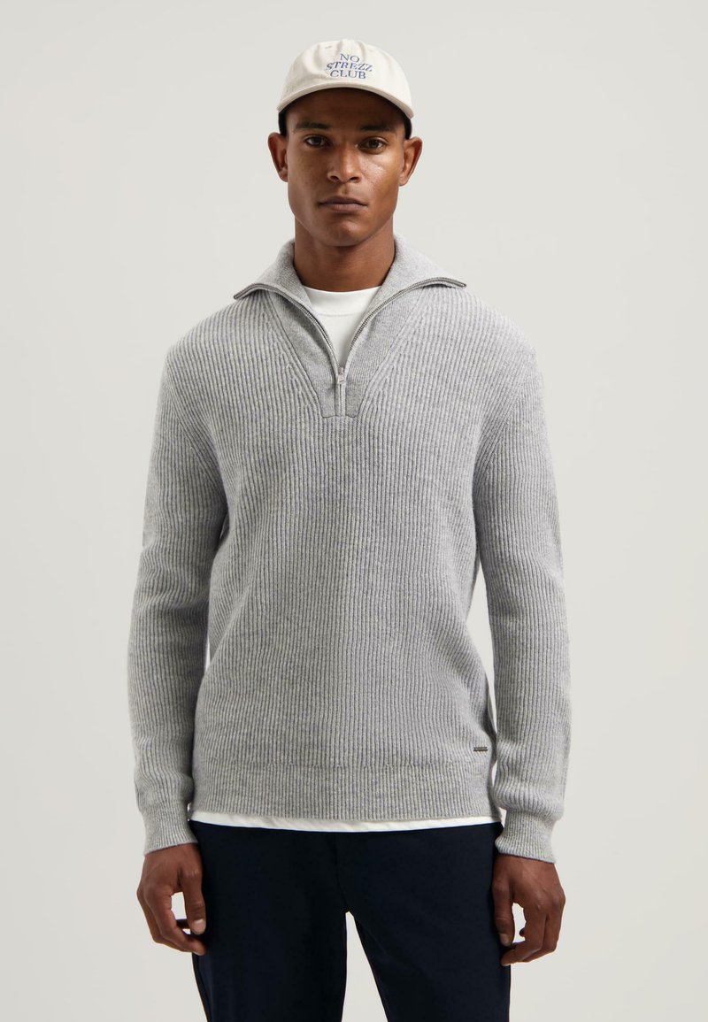 Man in een lichtgrijze geribbelde sweater met kwartrits, een wit T-shirt eronder, marineblauwe broek en een witte pet met blauwe tekst "NO STREZZ CLUB".