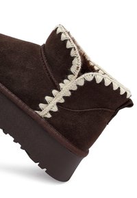 Bruine suède enkellaars met een zachte binnenkant, voorzien van crèmekleurige decoratieve stiksels langs de rand en een stevige rubberen zool.