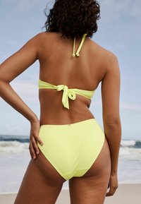 Neon žuti bikini s halter vratnim dizajnom i zatvaranjem na vezanje. Visoki pojas na donjem dijelu ima glatku teksturu i minimalne detalje od hardvera.