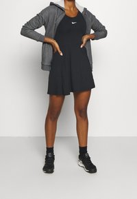 Svart sportklänning med figursydd överdel och utsvängd kjol, kombinerad med en grå zip-up huvtröja. Svarta sneakers och ankelsockor kompletterar looken.