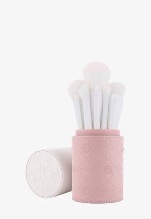 Ensemble de pinceaux de maquillage blancs avec têtes de différentes tailles dans un porte-pinceaux cylindrique texturé rose, avec un couvercle blanc assorti posé à côté.