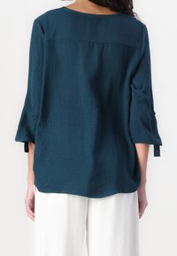 Losse, donkergroen blouse in gestructureerde stof met driekwartmouwen en knoopaccenten op de manchetten. Ronde hals aan de achterkant.