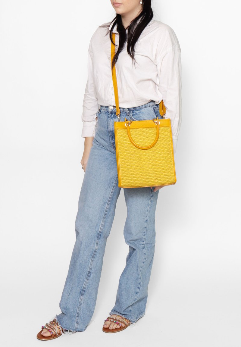 Borsa gialla con superficie texturizzata, forma rettangolare, brevi manici circolari e una lunga tracolla regolabile. Indossata con jeans azzurri.