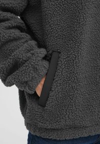 Gray fleece pullover featuring a textured surface, a black sleeve trim, and side pockets. The fabric is soft and fuzzy in appearance.