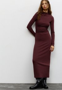 Abito lungo bordeaux a maniche lunghe con scollo alto e silhouette aderente, realizzato in tessuto morbido. Abbinato a stivali neri al ginocchio.