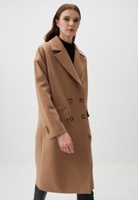 Cappotto doppiopetto color cammello con colletto a punte, caratterizzato da grandi bottoni e due tasche laterali. Realizzato in un tessuto strutturato e caldo.