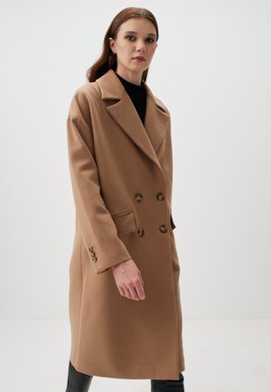 Cappotto doppiopetto color cammello con colletto a punte, caratterizzato da grandi bottoni e due tasche laterali. Realizzato in un tessuto strutturato e caldo.
