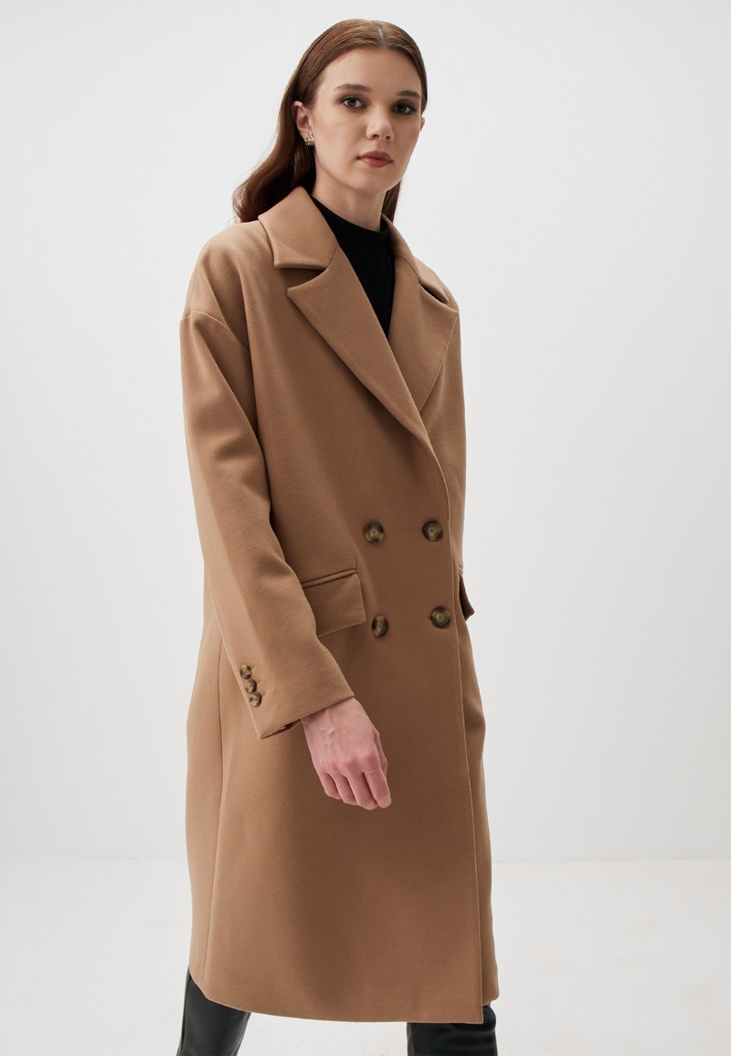 Cappotto doppiopetto color cammello con colletto a punte, caratterizzato da grandi bottoni e due tasche laterali. Realizzato in un tessuto strutturato e caldo.